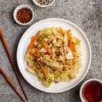 A plate of carrot cabbage stir-fry with chopsticks, tea, chili flakes, and sesame seeds beside.