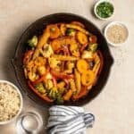 A pan of stir-fry vegetables with rice, water, green onion, and sesame seeds beside.