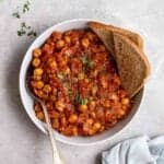 A bowl of tomato chickpeas with spoon and toasted bread beside.