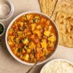 A bowl of vegetable madras curry served with spoon with rice and chapati beside.