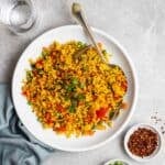 A plate of Indian fried rice served with spoon with chili flakes, green onion, and water beside.