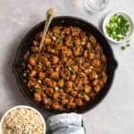 A pan of soya chunk manchurian served with spoon with green onion, glass of water, and bowl of rice beside.