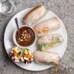 A plate of spring rolls served with a dish of peanut sauce with glass of water beside.