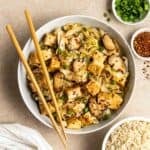 A bowl of cabbage tofu stir-fry served with chopsticks with bowl of rice, green onion, and chili flakes beside.