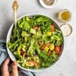 Holding a bowl of avocado lettuce salad served with fork with dressing and black pepper beside.