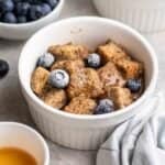 Mug french toast topped with bleberries with bowl of blueberries, maple syrup, and a blue teacloth beside.