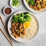 Orange tofu served with rice and broccoli on a plate with chopsticks beside.