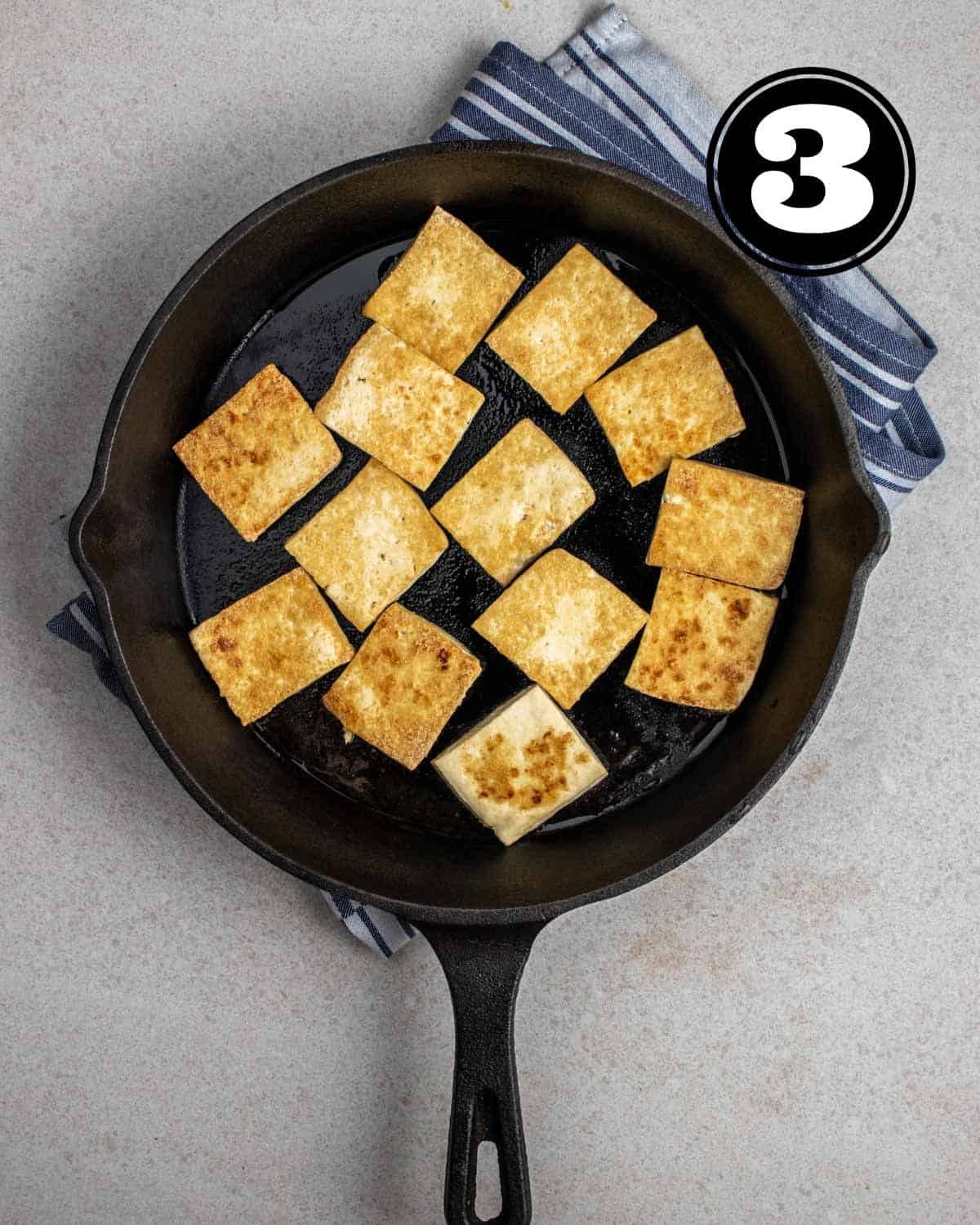 Pan-fried tofu in a cast iron skillet on a folded white and blue striped teacloth.