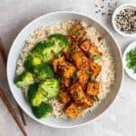 A plate containing rice, gochujang tofu, and broccoli with chopsticks, sesame seeds, and green onion beside.