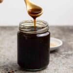 Spooning out some teriyaki sauce from a jar with a small plate of sesame seeds in the background.