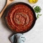 A pan of vegan marinara sauce with a wooden stirrer, olive oil, and chopped basil beside.