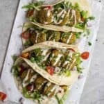 4 tacos arranged on a parchment paper drizzled with tahini sauce.