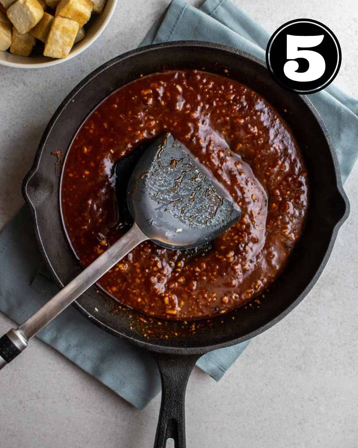Gochujang sauce in a cast iron skillet with a spatula beside on a blue folded teacloth.