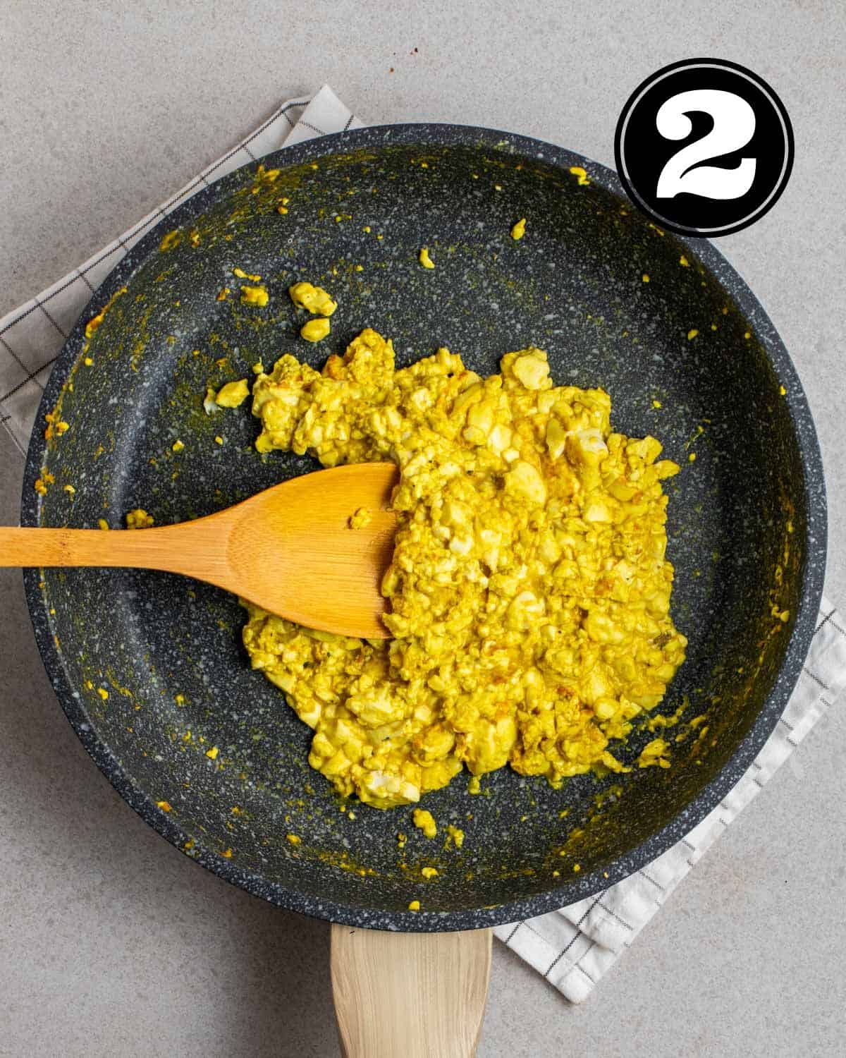 Cooked silken tofu scramble in a pan with a wooden spatula beside on a white folded teacloth.
