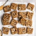 Slices of flapjacks arranged on a parchment paper with spoonful of peanut butter and bowl of oats beside.
