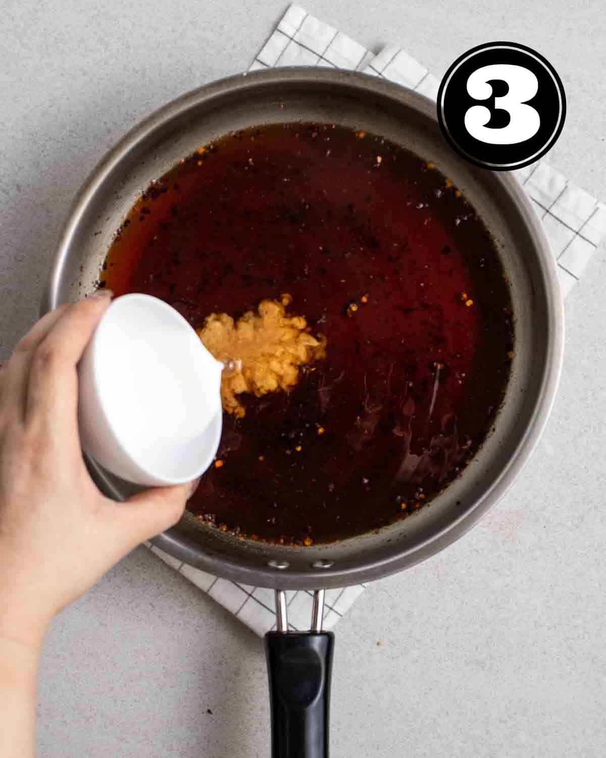 Pouring cornstarch into a saucepan with teriyaki sauce on a white folded teacloth.