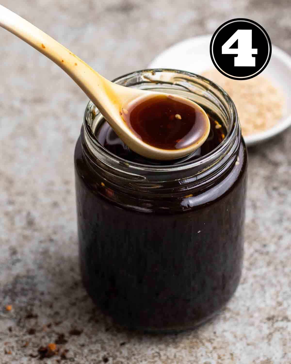 A jar of spicy teriyaki sauce with a spoon on top with a plate of sesame seeds in the background.