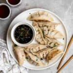 A plate of dumplings served with soy sauce with a pair of chopsticks and 2 cups of tea beside.