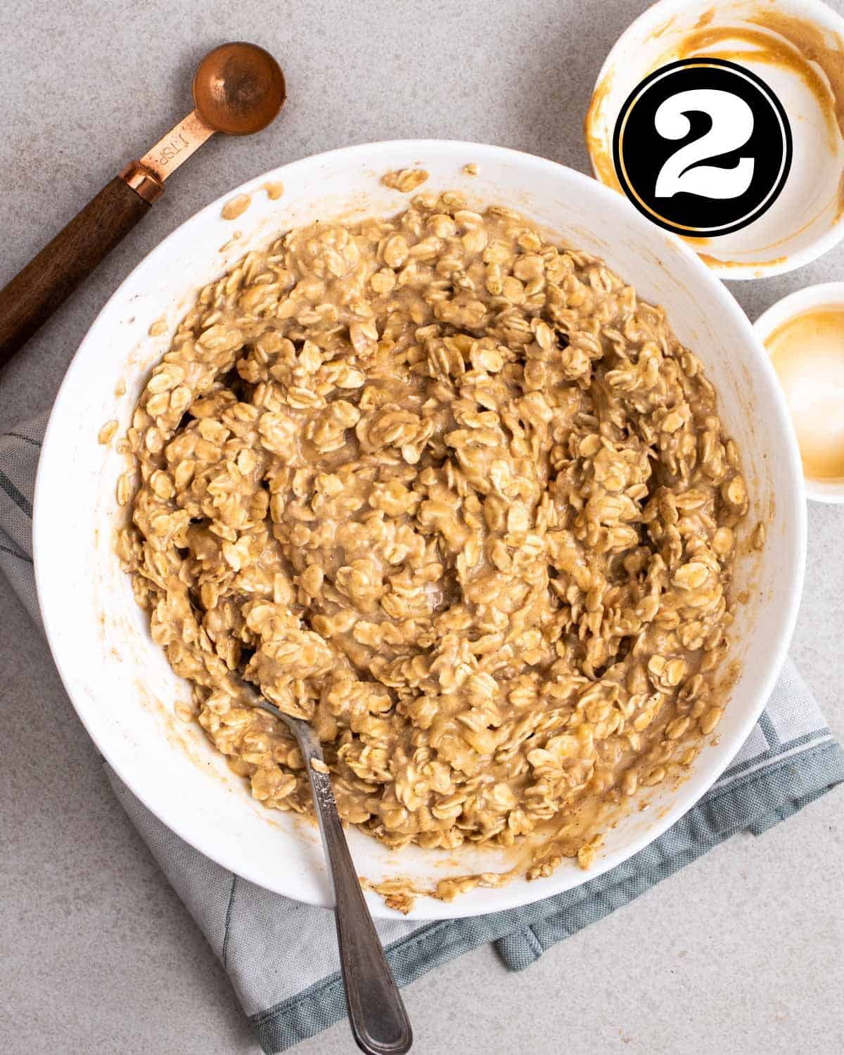 Banana flapjack batter in a white bowl with a fork, 2 ramekins, and a teaspoon beside.