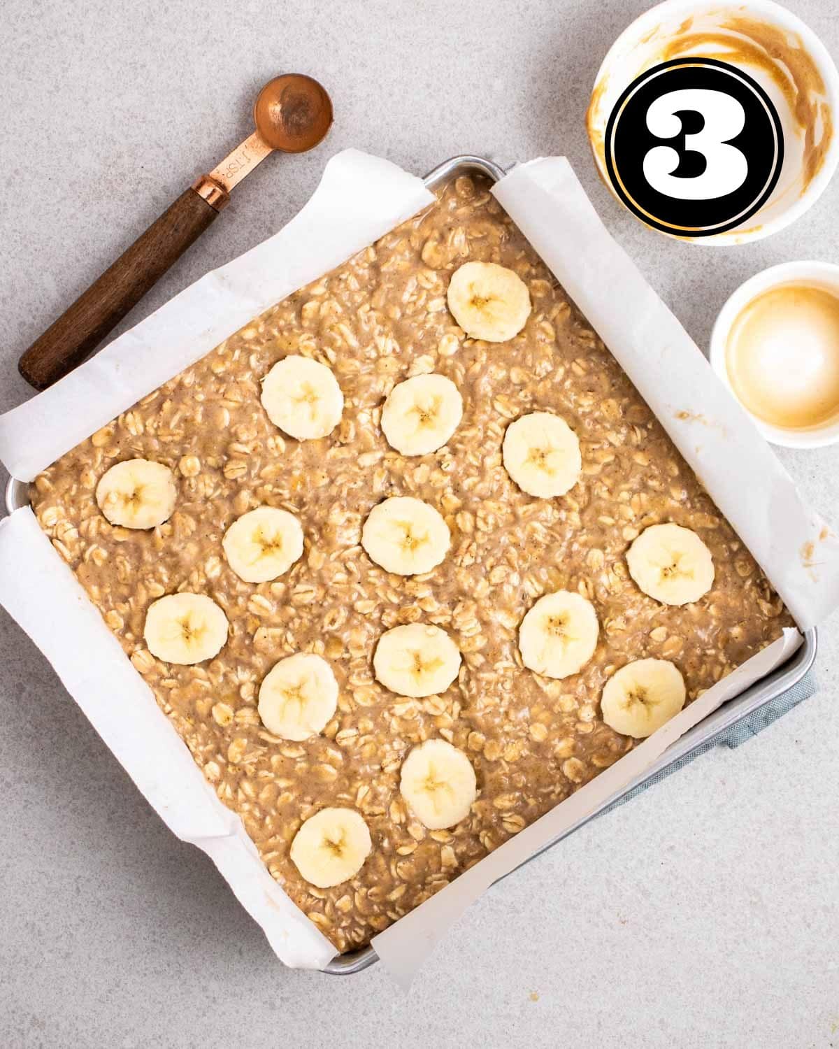 Unbaked banana flapjack in a pan lined with parchment paper and topped with banana slices.