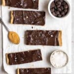 Protein bars arranged on a baking paper wirg spoonful of peanut butter, chocolate chips and sea salt beside.