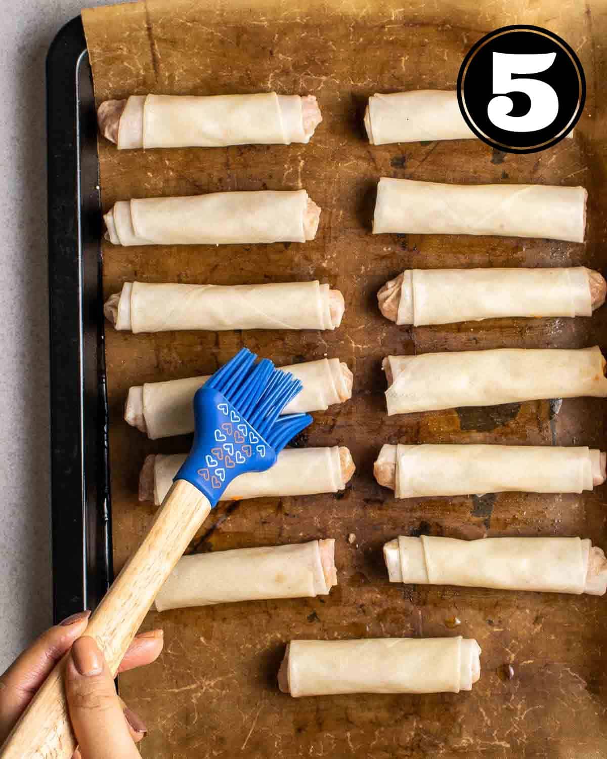 Brushing on some oil on vegan lumpia arranged on a lined baking sheet.