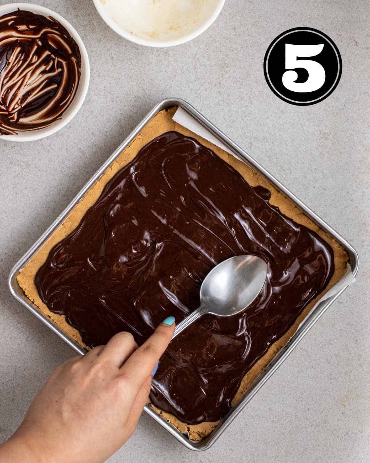 Spreading melted chocolate onto protein bar dough pressed in a square pan.