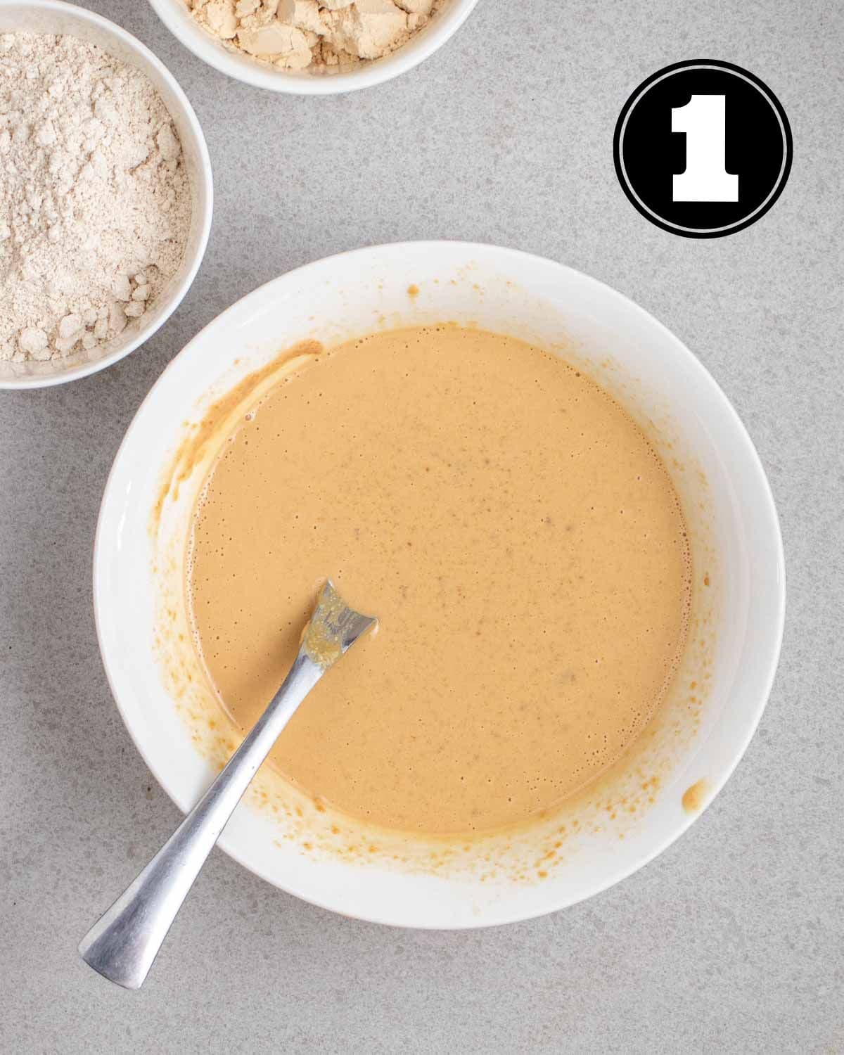 Wet ingredients for protein bars in a white bowl with a spoon beside.