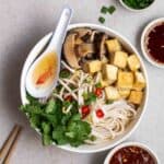 A bowl of pho served with spoon with chopsticks, green onion, and condiments beside.