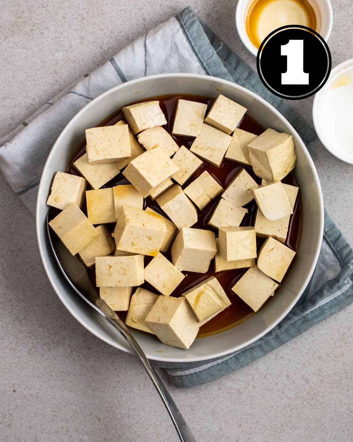 Marinating tofu cubes in adobo sauce in a deep plate with a spoon beside.