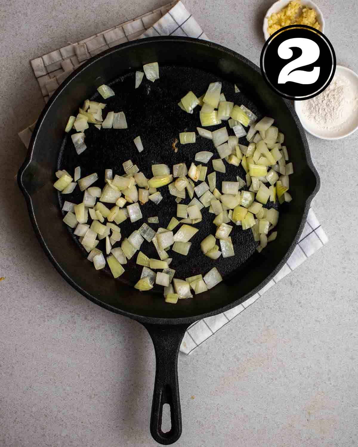 Cooked onion in a cast iron skillet on a folded white teacloth with flour and minced garlic beside.