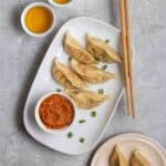 A plate of momos served with chutney and chopsticks with 2 cups of tea beside.