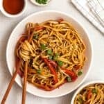 A plate of noodles served with chopsticks with another bowl of noodles, tea, and chopped green onion beside.