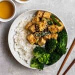 A plate of rice served with tofu and broccoli with a pair of chopsticks and 2 cups of tea beside.