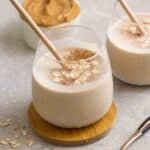 2 glasses of peanut butter smoothie served with straws with peanut butter in the background.