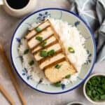 Sliced tofu on a bowl of rice with chopsticks, chopped green onion, and soy sauce beside.