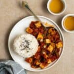 A plate of rice and tomato sauce tofu served with a spoon with 2 cups of tea beside.