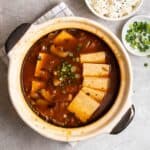 A claypot of kimchi soup on a checkered white teacloth with rice and chopped green onion beside.