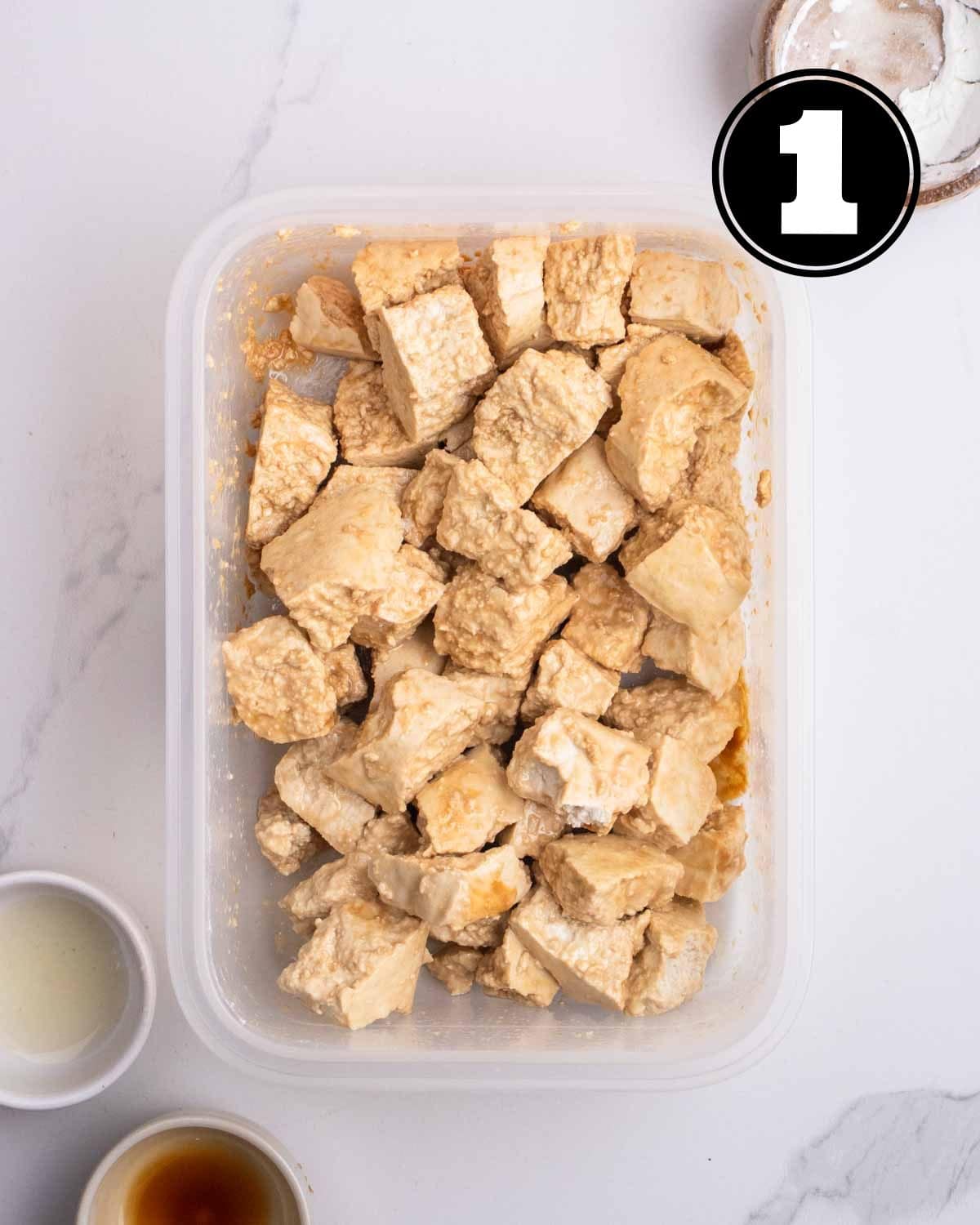 Tofu coated in cornstarch and soy sauce in a container with 2 sauce dishes beside.