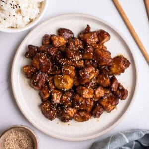A plate of hoisin tofu with bowl of rice, pair of chopsticks, and dish of sesame oil in the background.