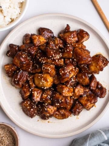 A plate of hoisin tofu with bowl of rice, pair of chopsticks, and dish of sesame oil in the background.