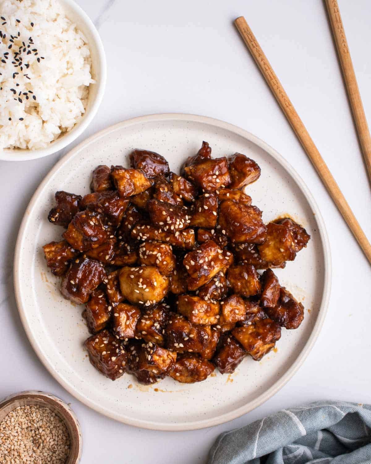 A plate of hoisin tofu with a bowl of rice, pair of chopsticks, and sesame seeds beside.