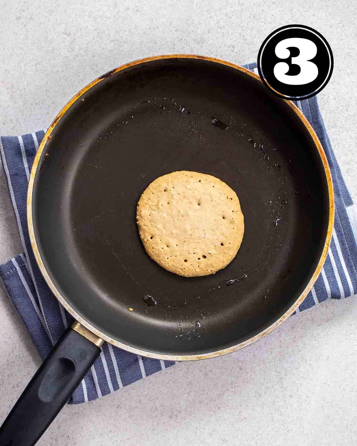 A partially cooked pancake in a non-stick pan on a blue and white striped teacloth.