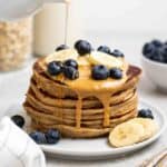 Pouring maple syrup over a stack of pancakes with oats, milk, and bowls of blueberries in the background.