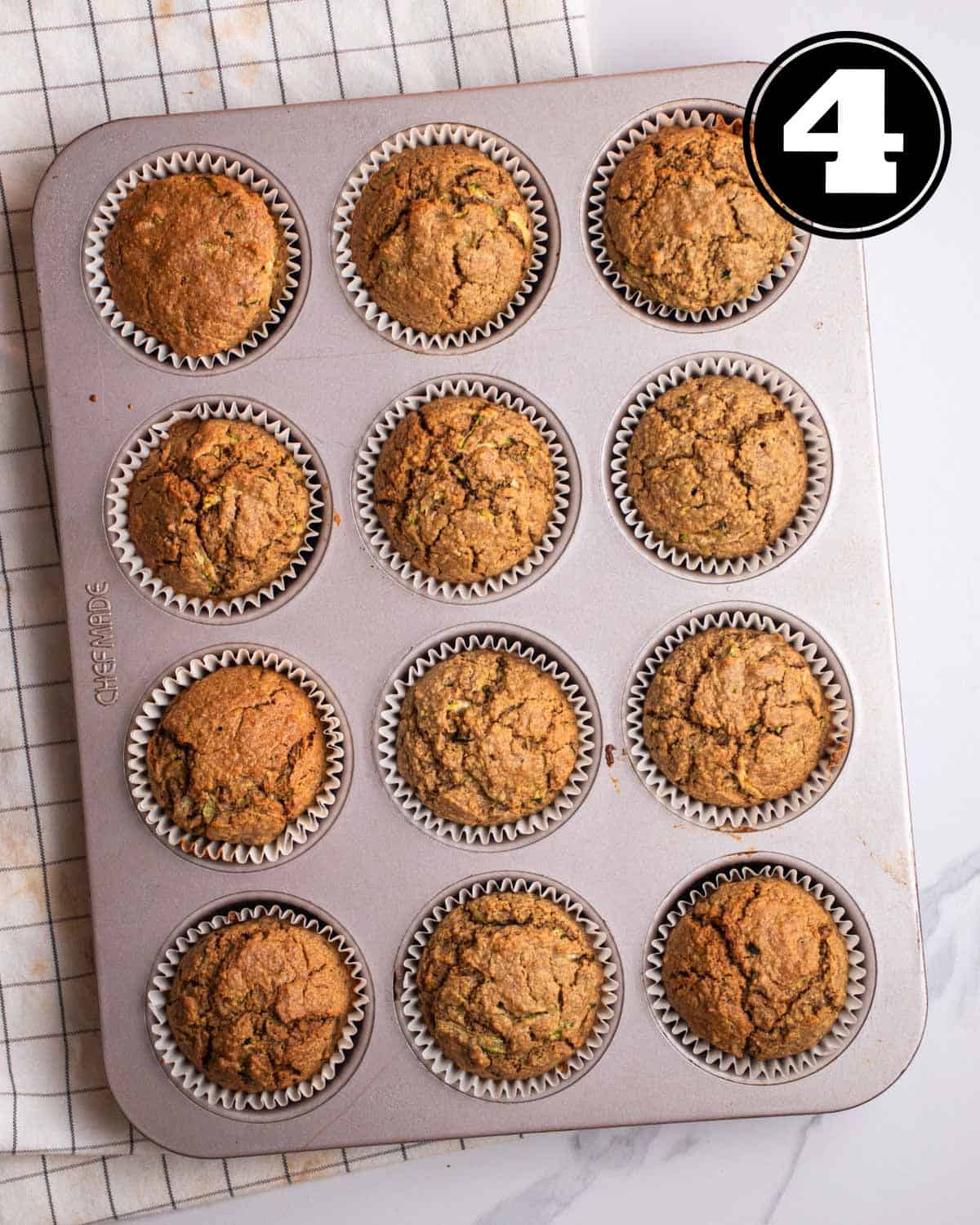 Baked muffins in a muffin tray on a checkered white teacloth.