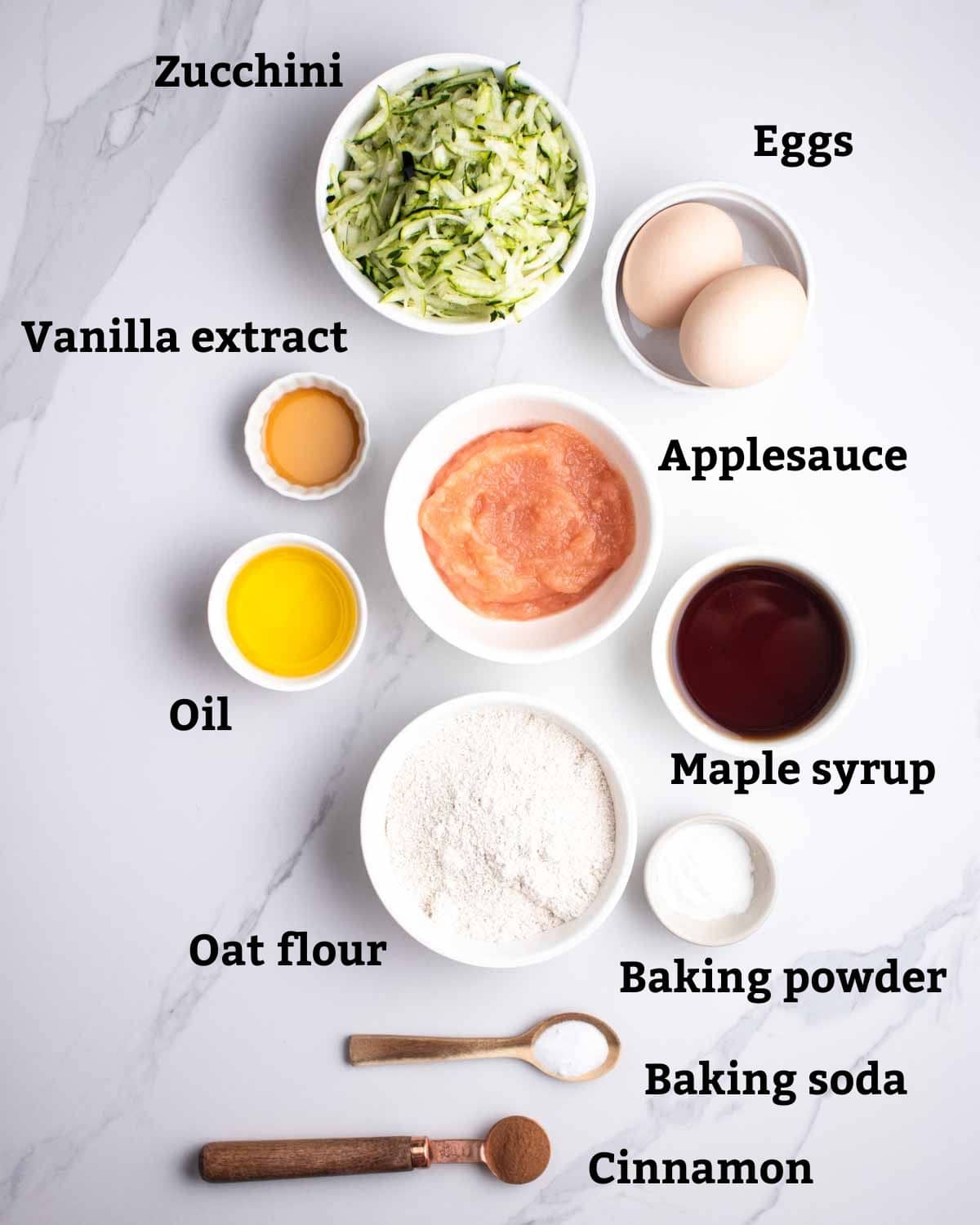 Ingredients needed such as oat flour, maple syrup, eggs, zucchini, and applesauce arranged on a white background.