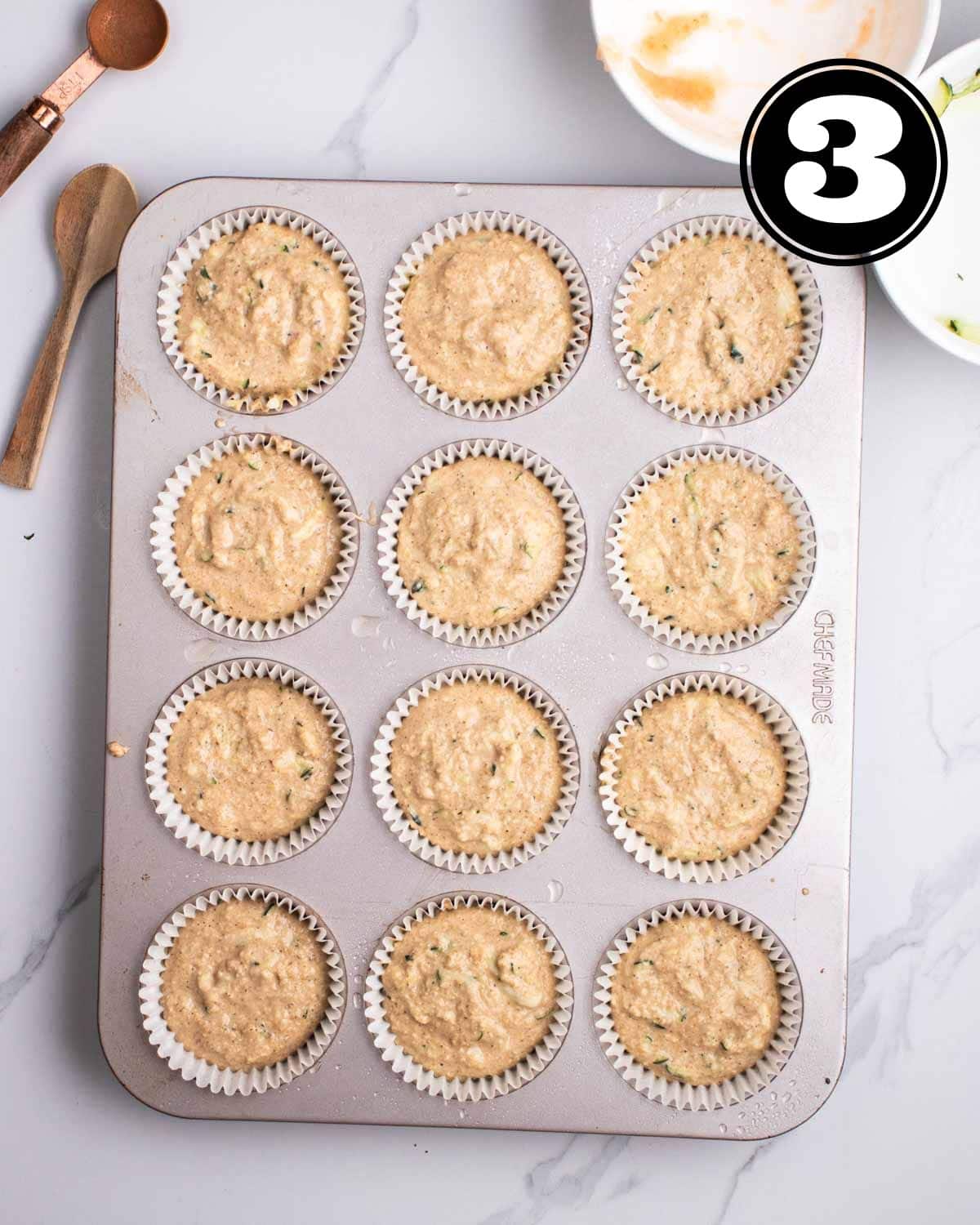 Unbaked applesauce zucchini muffins in a muffin tray with 2 teaspoons and 2 empty ramekins beside.