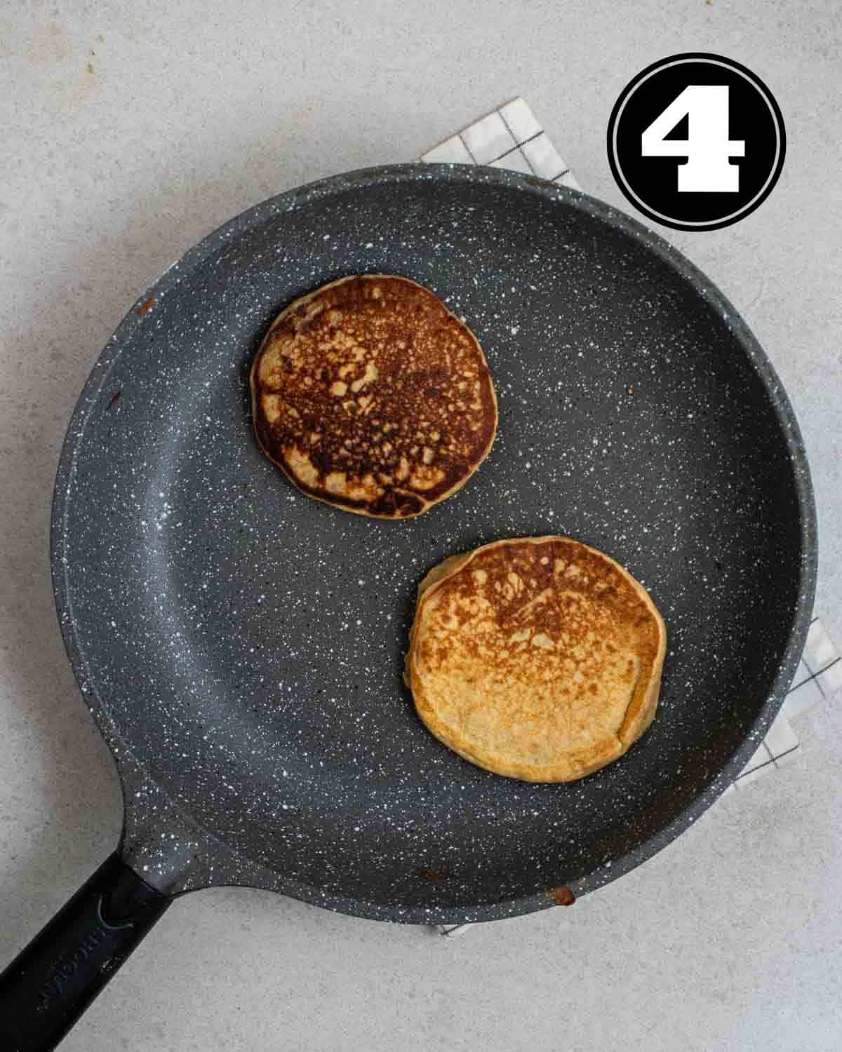 2 pieces of cooked pancakes in a non-stick pan on a white folded teacloth.