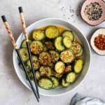 A plate of cucumber salad served with chopsticks with sesame seeds and chili flakes beside.