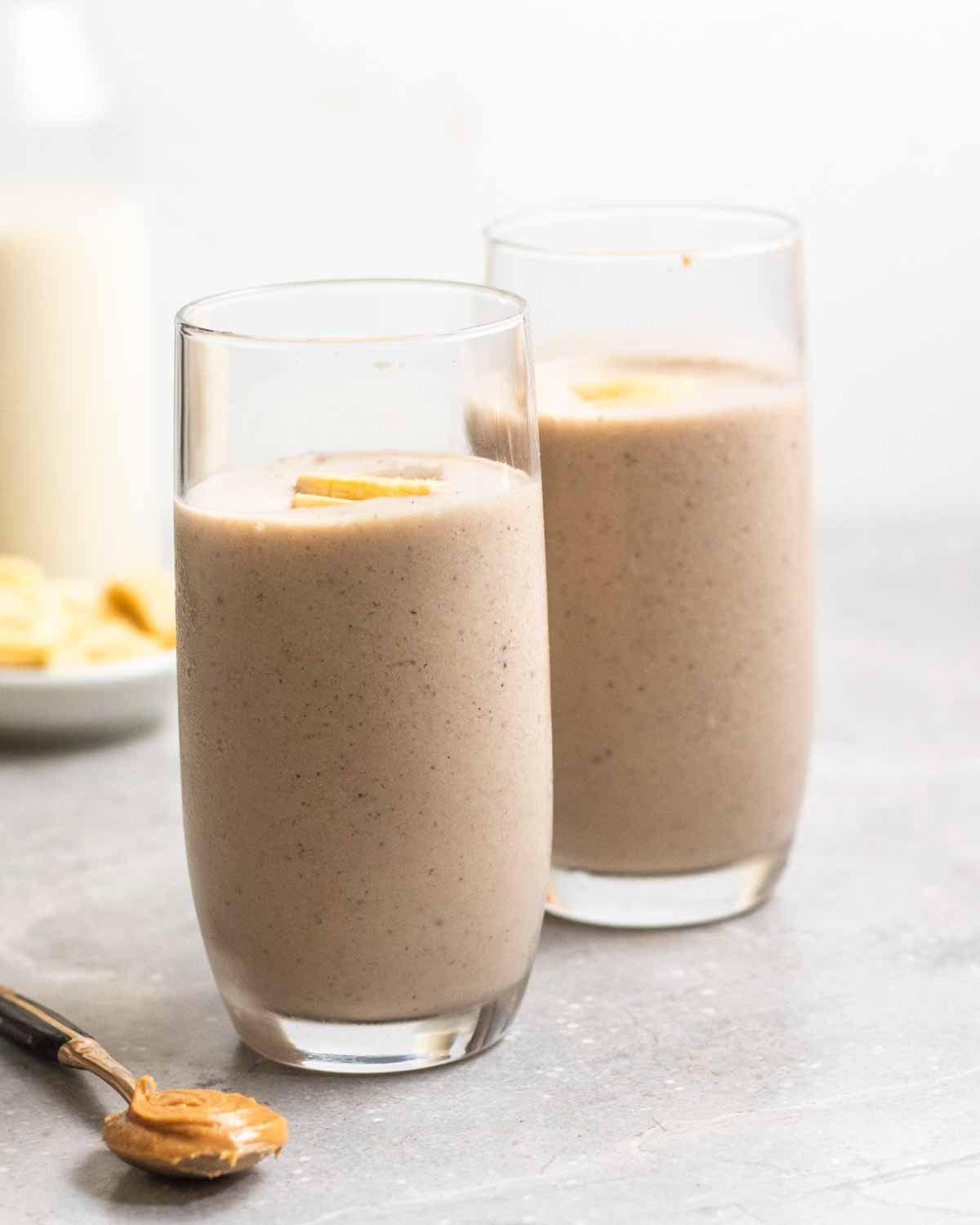 2 glasses of tofu smoothie with a teaspoon of peanut butter, banana slices, and jug of soy milk in the background.